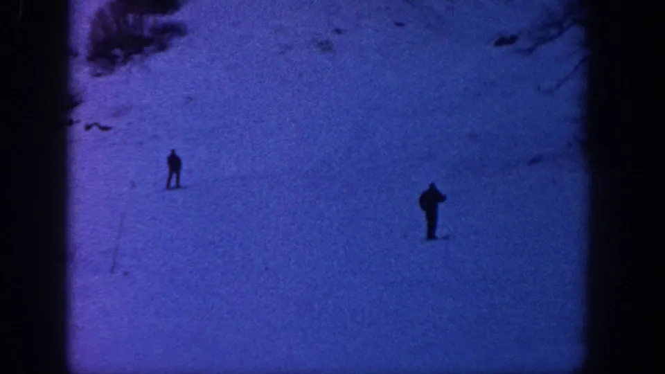 Two distant skiers are silhouetted on a broad Aspen snow slope, with sparse brush and dark frame edges from the 8mm scan.