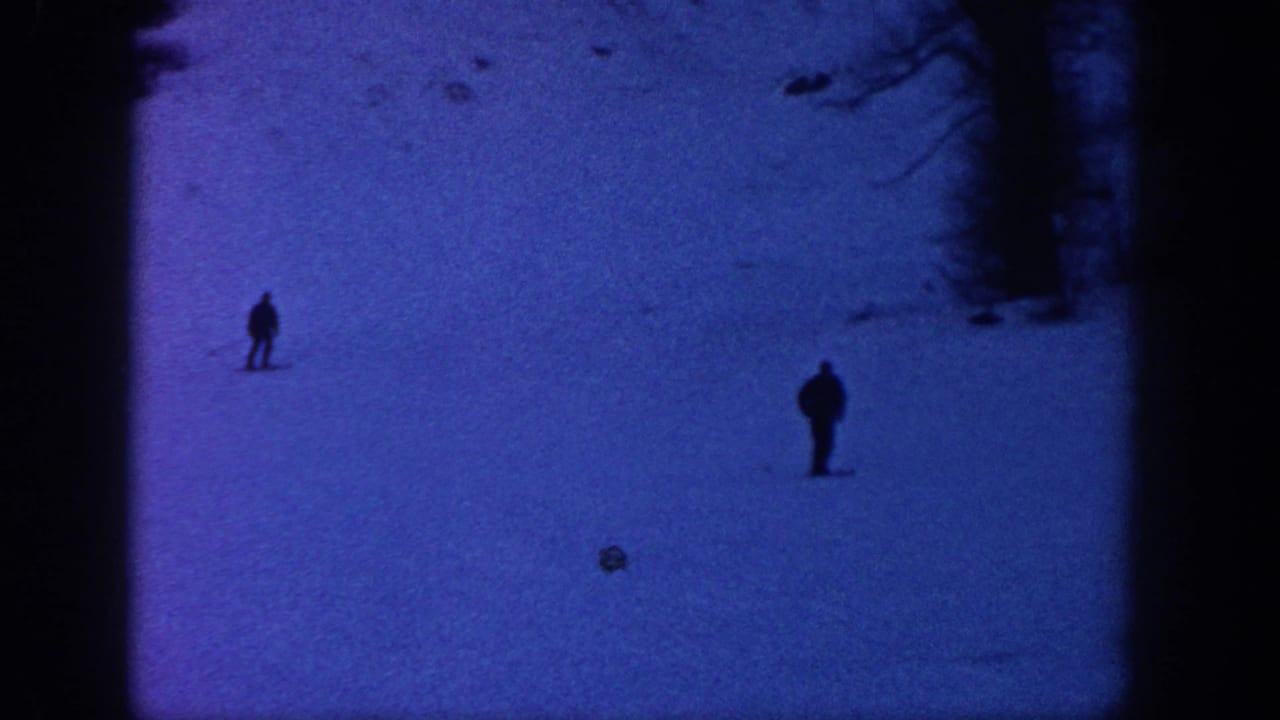 1961 Aspen home-movie frame showing two distant skiers silhouetted on a blue snowy slope below bare trees.