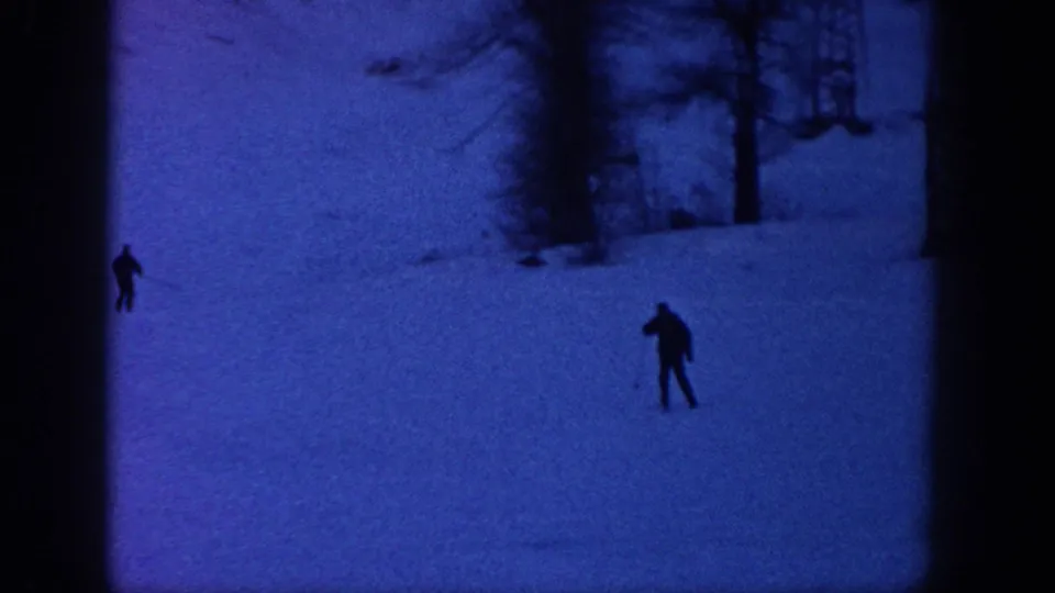 1961 Aspen ski footage frame with two dark skier silhouettes crossing a blue snowfield near trees and a lift support.