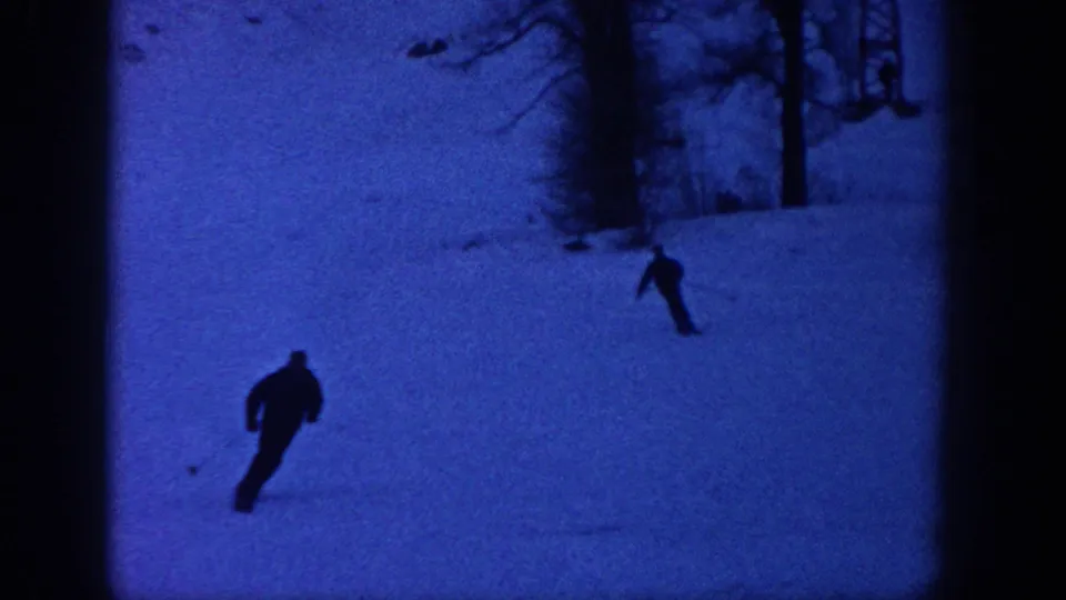 1961 Aspen home-movie frame showing a foreground skier turning across the slope with another skier higher near the trees.