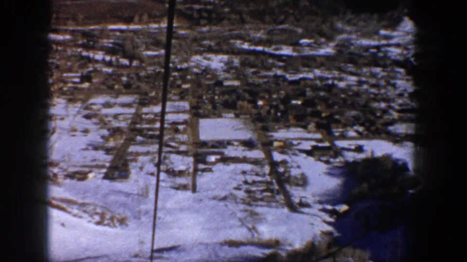 A snow-covered townscape is seen from above in soft, grainy 8mm footage, with roads, buildings, and dark film-edge shadowing.