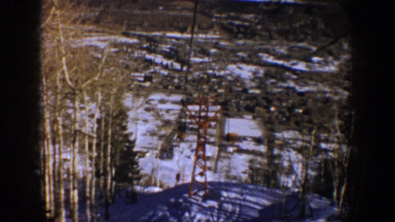 1961 Aspen home-movie frame showing a red ski-lift tower on a snowy slope with bare aspen trees, evergreens, and distant buildings.