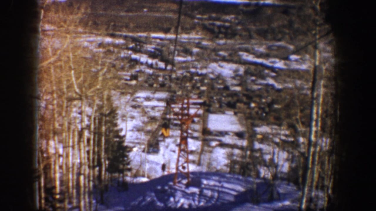 Red ski-lift tower and cable above a snowy slope with trees and a small moving figure.