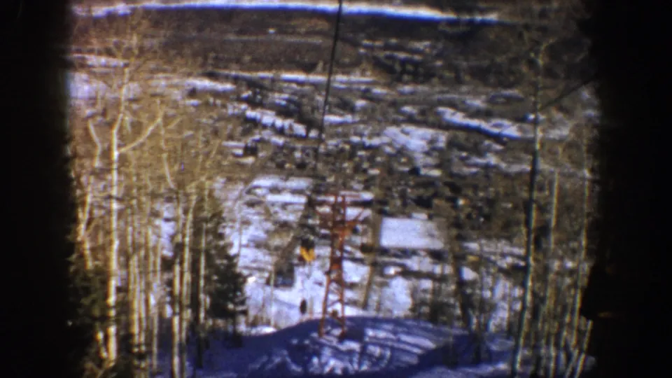 Aspen 1961 8mm frame showing a red ski-lift tower and cable across a snowy wooded slope with distant buildings.