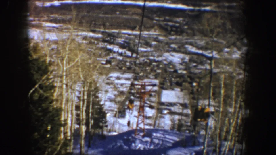 1961 Aspen frame showing the lift tower and cable on a snowy wooded slope, with a small distant chair or rider form visible.
