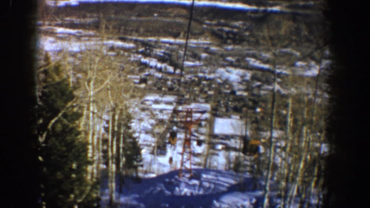 1961 Aspen home-movie frame showing a patchy snowy valley, lift cables, bare aspen trees, evergreens, and distant buildings.
