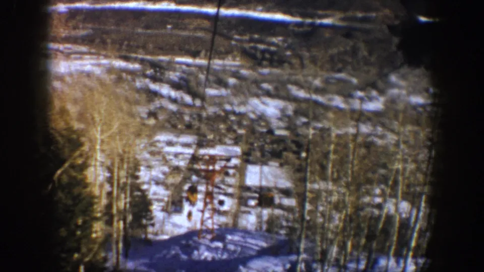 1961 Aspen frame with lift cables and a small lift structure above melting snow, bare trees, evergreens, and valley buildings.