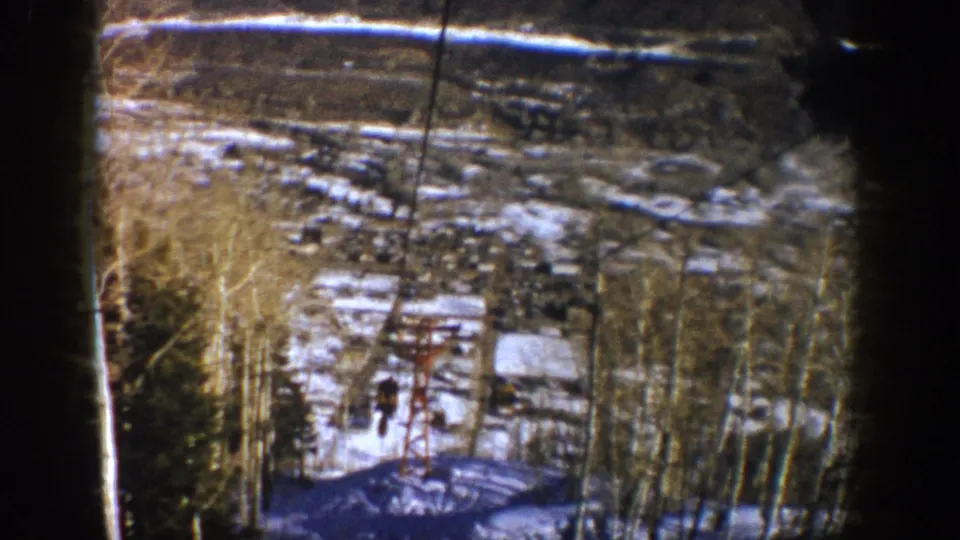 1961 Aspen home-movie view of a ski-lift line over a thawing slope, with bare aspen trunks, buildings, and distant terrain.