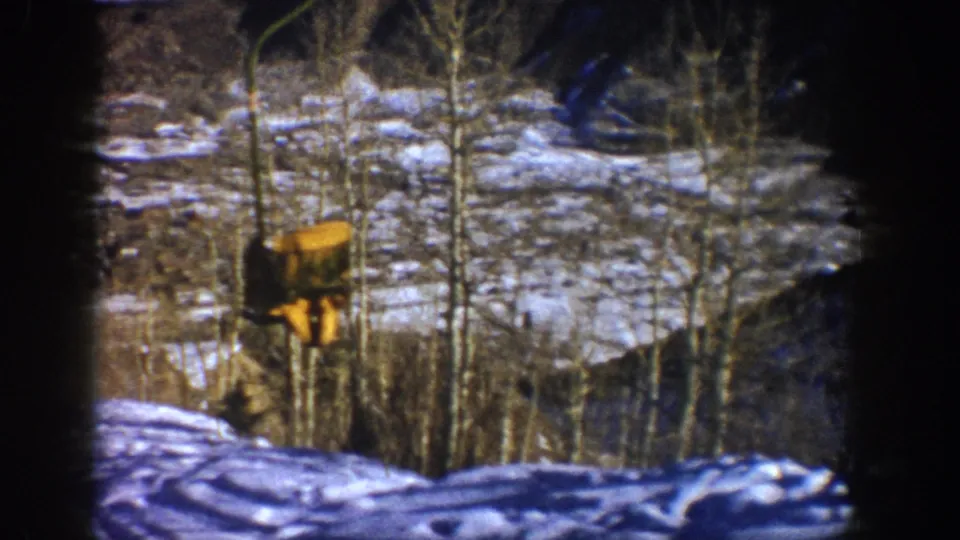 1961 Aspen frame showing a yellow lift car moving along cables above patchy snow, bare aspen trees, and a wooded mountain slope.