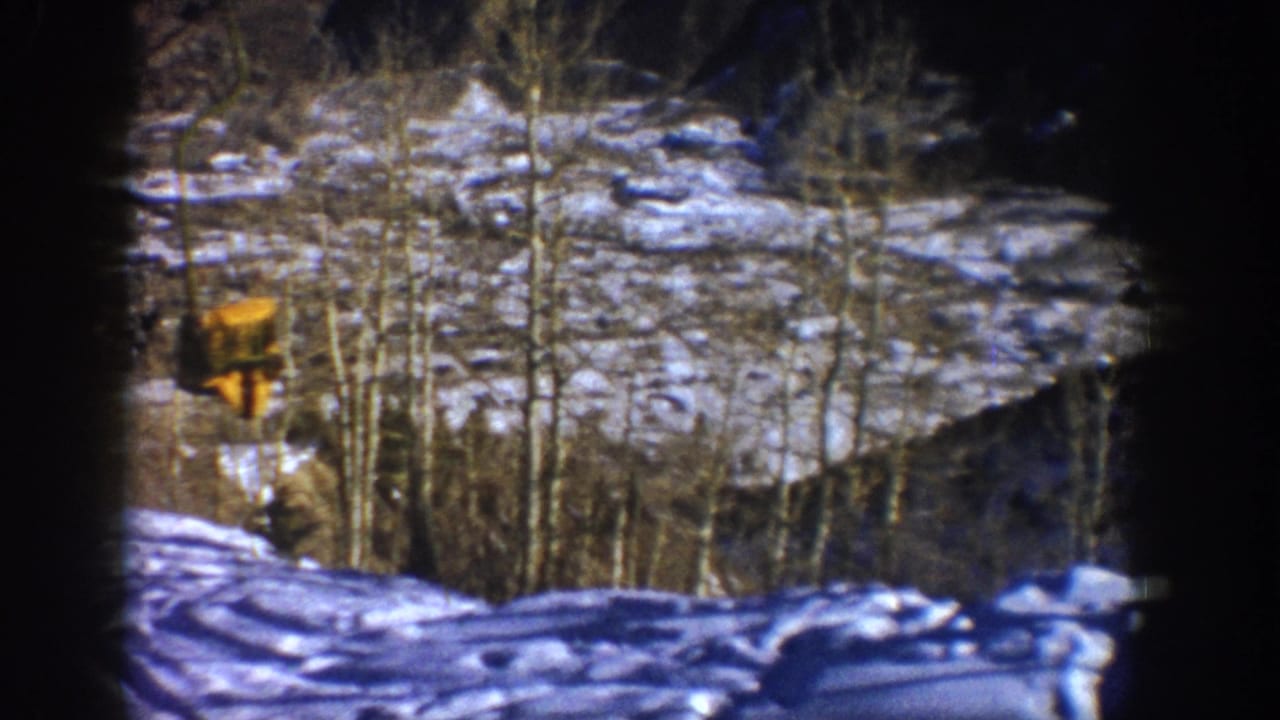 Aspen, Colorado winter home-movie frame from 1961 showing snow, bare aspen trees, and a distant yellow structure through dark film edges.