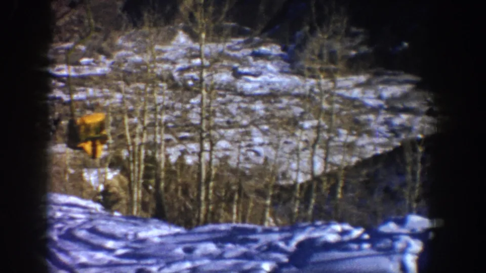 Aspen, Colorado winter home-movie frame from 1961 showing snow, bare aspen trees, and a distant yellow structure through dark film edges.