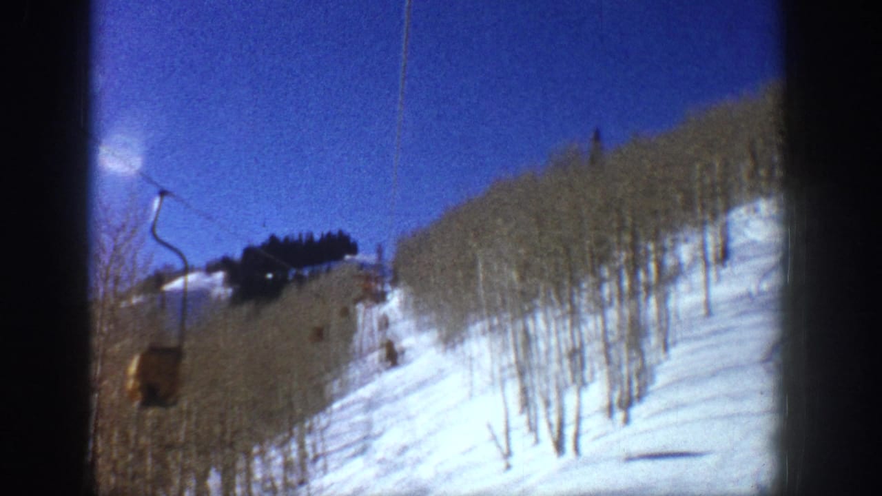 1961 Aspen frame with a small yellow ski lift cabin moving along a cable over a snowy hillside lined with bare aspen trees.