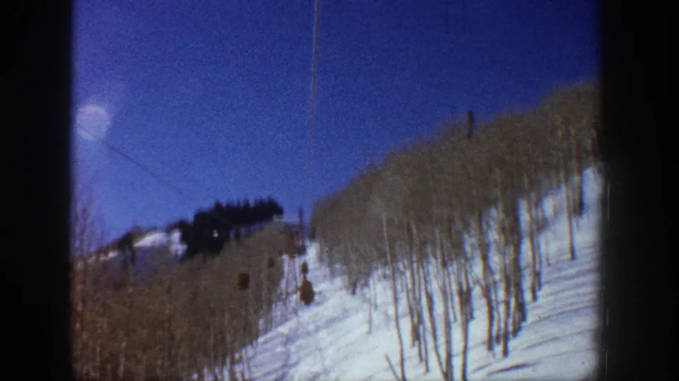 1961 Aspen 8mm frame showing lift cabins on a cable above a wooded snow slope, with blue sky and dark treeline near the ridge.