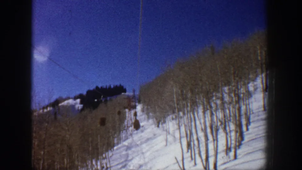 1961 Aspen frame with ski lift cabins, bare aspen trees, and a bright snow-covered slope seen through soft 8mm home-movie grain.