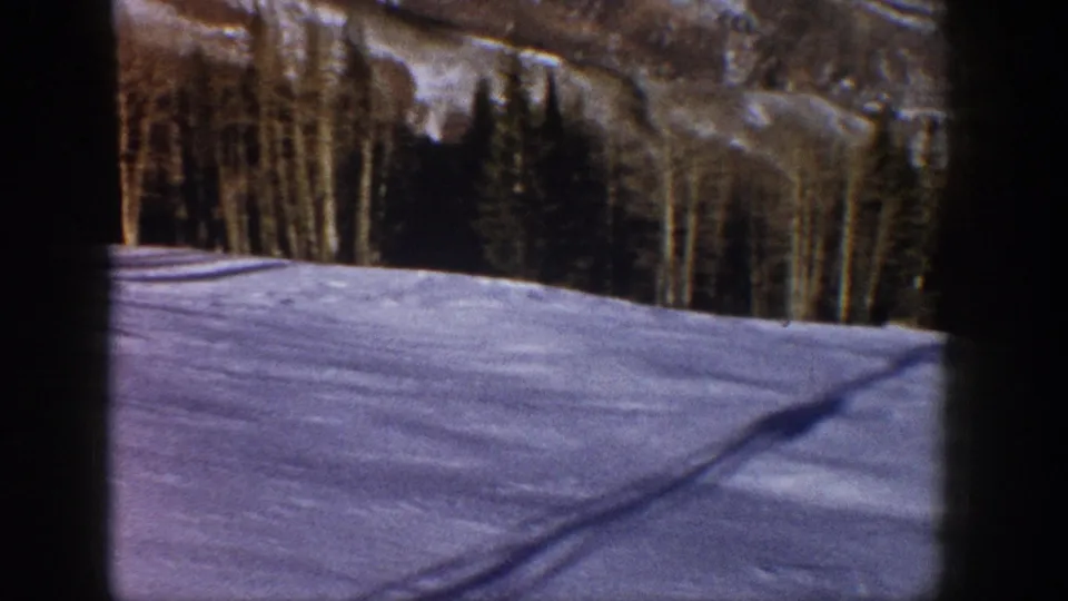 Passenger-view 8mm frame of snowy Aspen mountain terrain, with trees, shadows, and dark window edges.
