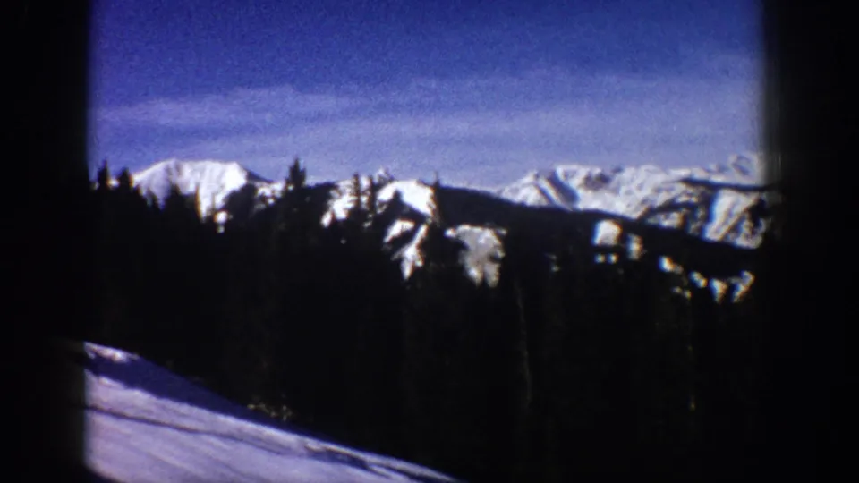 Distant snowy peaks near Aspen, Colorado appear beyond a dark evergreen forest in a 1961 8mm home-movie frame.