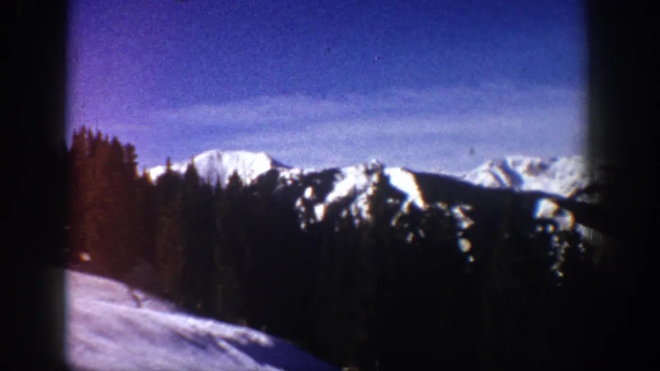 1961 Aspen, Colorado 8mm view of a snowy foreground slope, dense evergreen trees, and white mountain peaks in the distance.