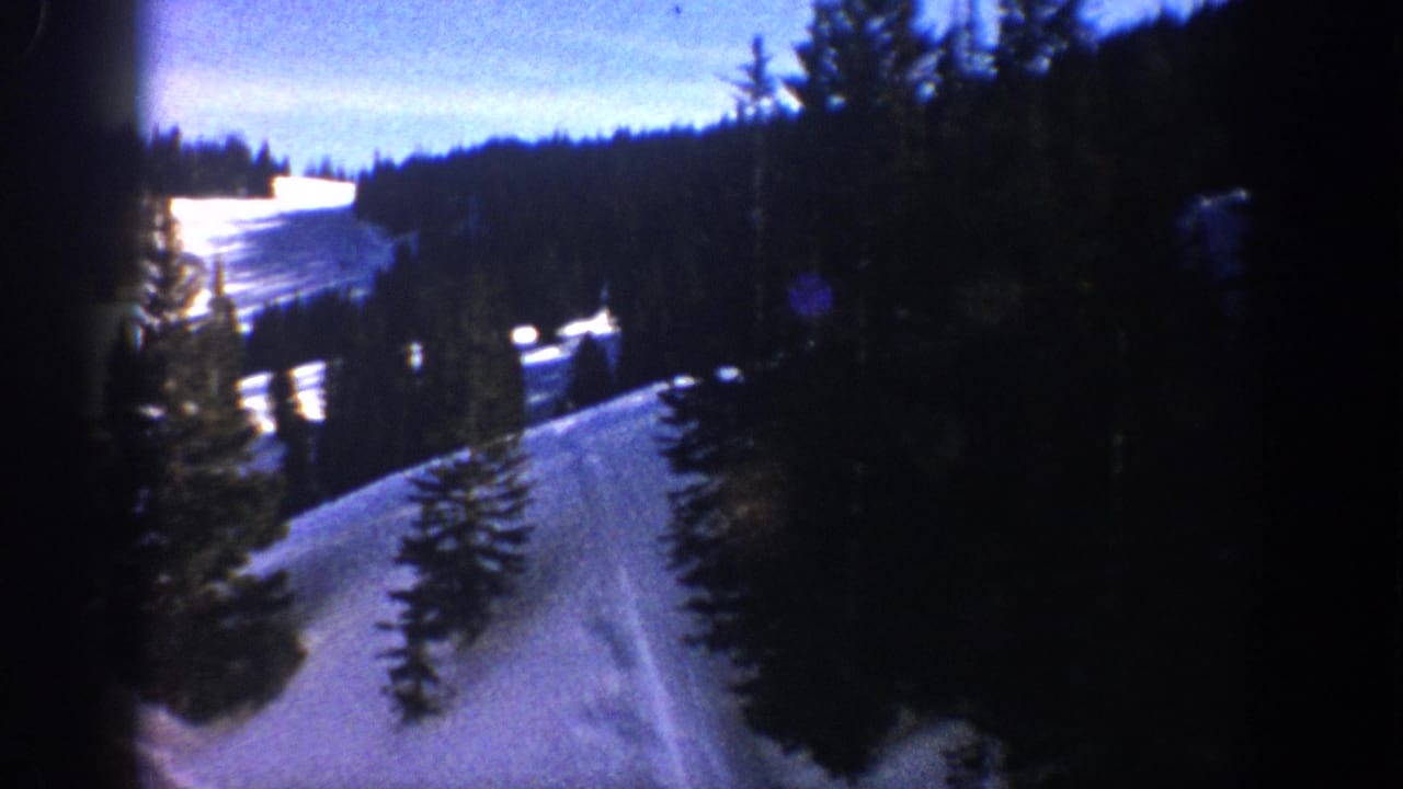 1961 Aspen, Colorado home-movie frame showing a snowy slope winding between dark evergreens at dusk.
