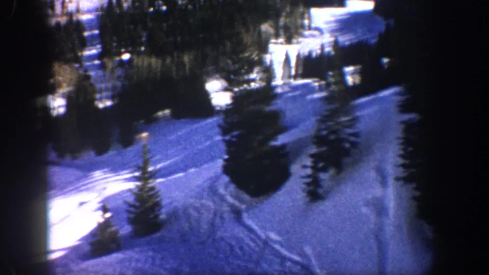 Vintage 8mm view of a snowy Aspen slope with evergreen trees, visible tracks, and low-light blue color cast.