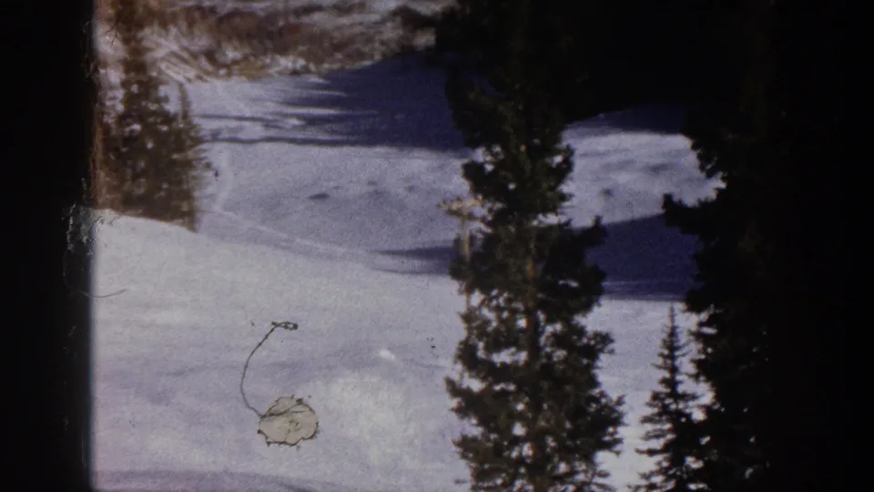 1961 Aspen home-movie frame showing a snowfield bordered by evergreens, faint slope tracks, and visible 8mm film surface marks.