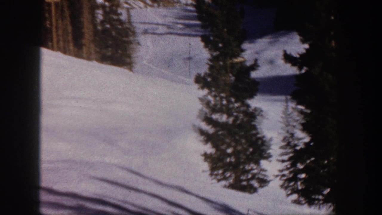 Aspen, Colorado 1961 home-movie frame showing a snowy ski slope bordered by evergreen trees, with chairlift equipment faintly visible in the distance.