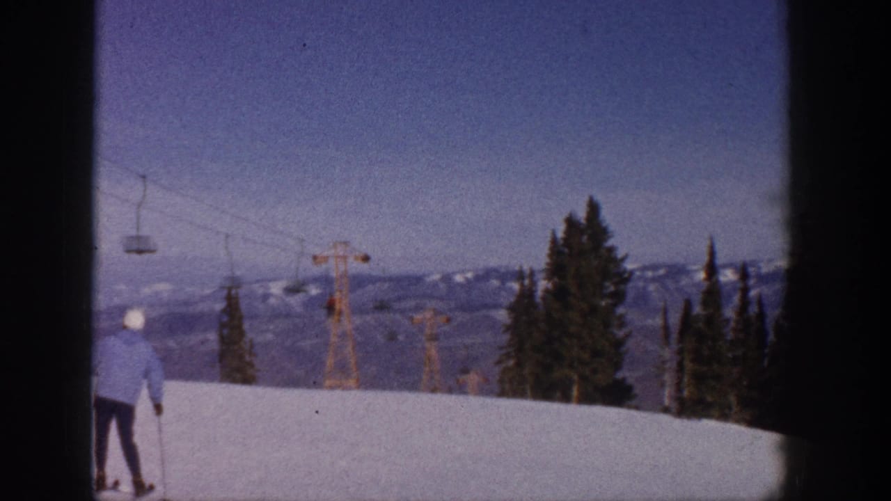 Skier on snowy slope with chairlift towers, evergreens, and mountains behind.