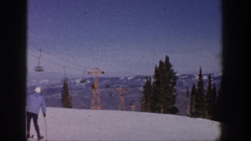 Aspen, Colorado 1961 frame with a skier in a light blue jacket on a snowy slope, chairlift towers and distant mountains behind.