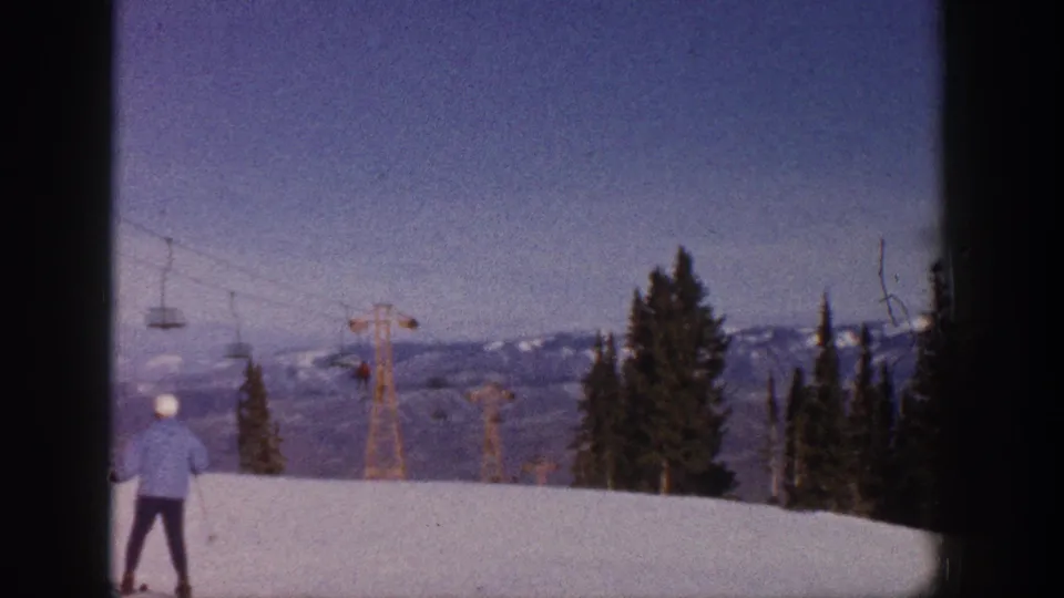 Aspen, Colorado 1961 home-movie frame showing a skier with poles on an open snow slope, with chairlift towers, evergreens, and mountains beyond.