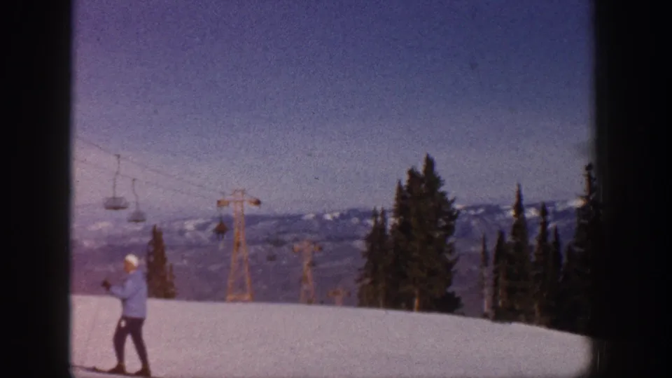 Aspen, Colorado 1961 frame showing a skier in a light blue jacket and white hat holding poles on a snowy slope near chairlift towers and evergreens.