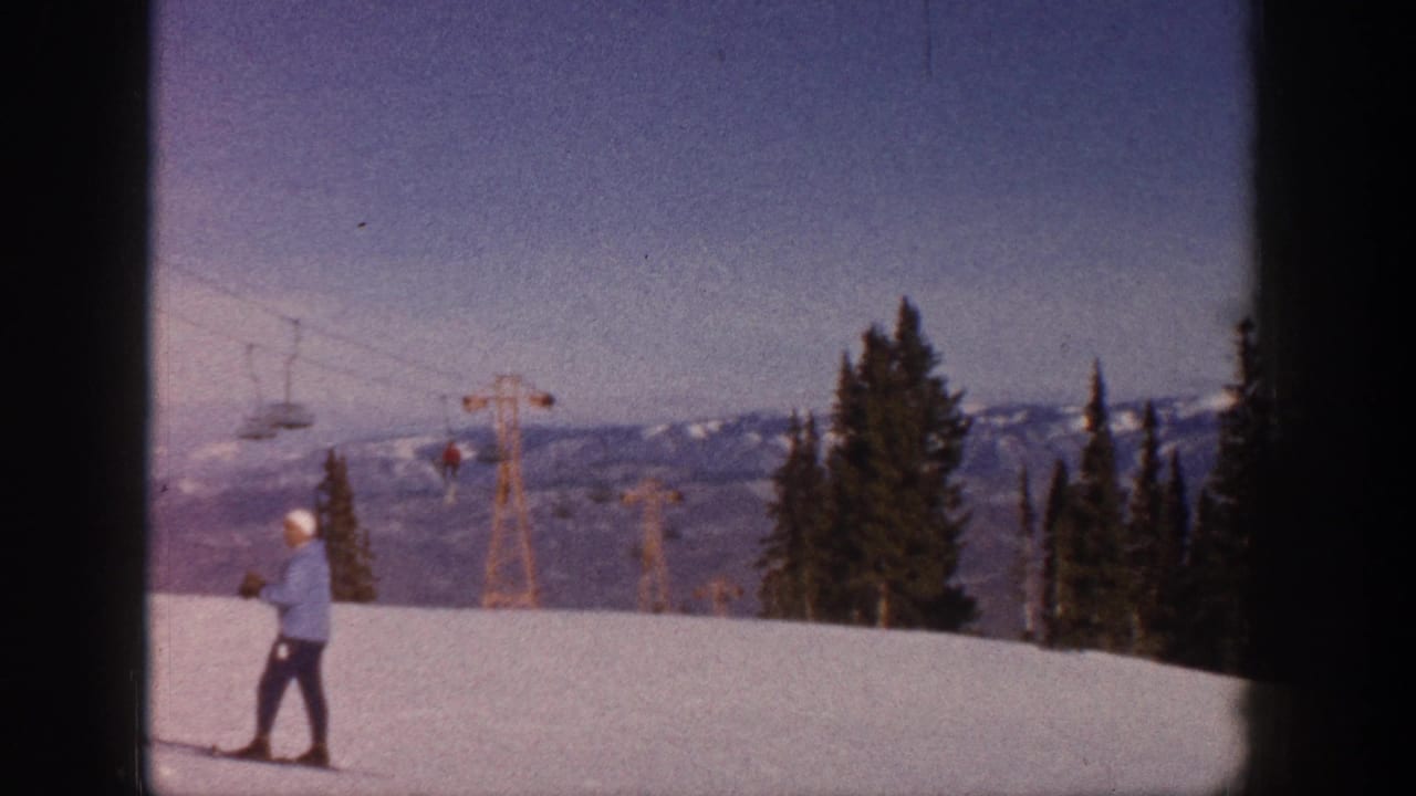 Skier on snow with chairlift cables, lift towers, trees, and mountain ridge in the background.