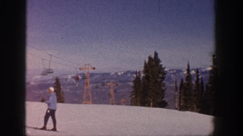 Aspen, Colorado 1961 frame of a skier on an open snowy slope beneath chairlift cables, with evergreens and mountains in view.
