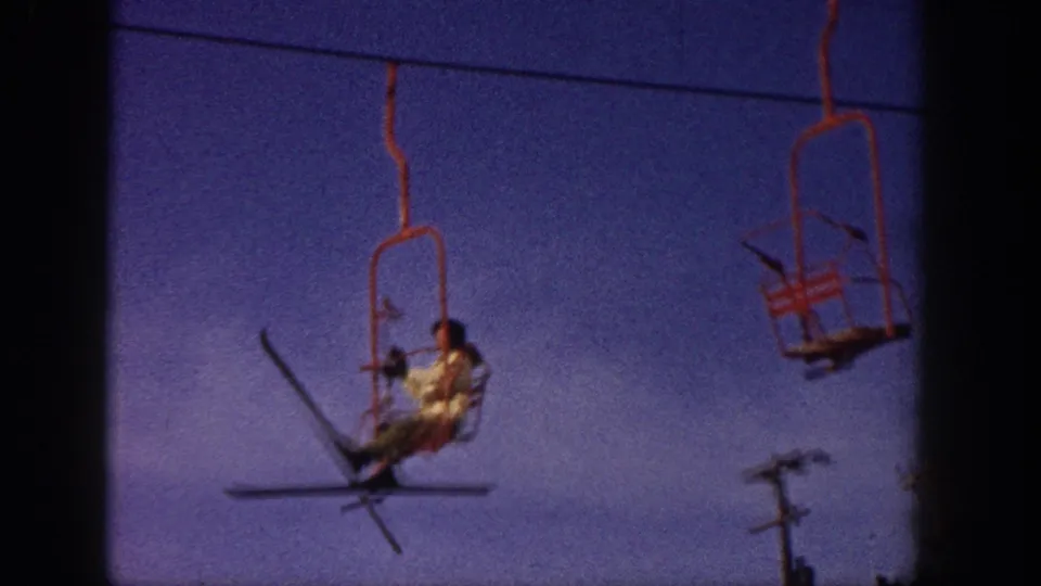 1961 Aspen chairlift view showing a skier seated on a red open lift chair, skis angled outward against a blue-purple sky.