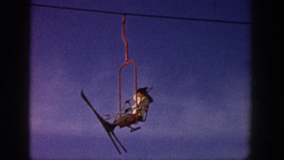 Aspen 1961 home-movie frame showing a skier riding a red chairlift, photographed close against the blue-purple winter sky.