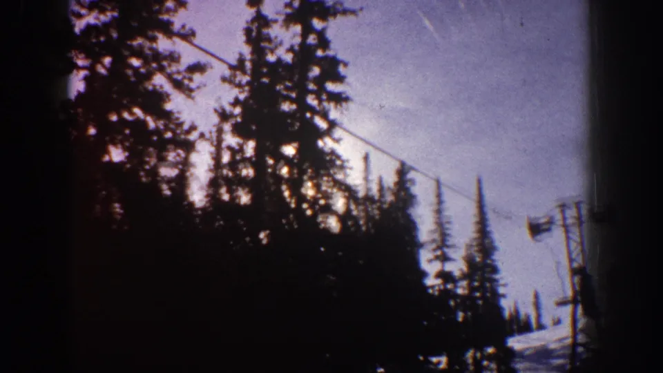 Handheld 8mm view of Aspen pines, ski lift infrastructure, and snow, with the sun partly hidden by the tree line.