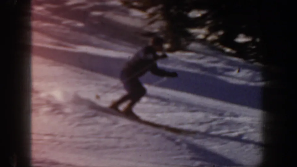 A distant skier moves downhill on a snow-covered Aspen slope, framed by shadowed trees and soft 8mm color blur.