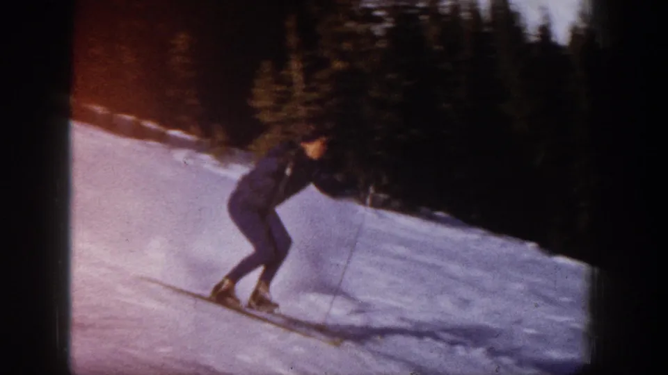 Home-movie frame from Aspen in 1961 showing a skier leaning into motion across snow below a dense tree line.