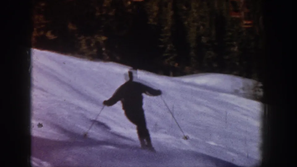 1961 Aspen 8mm frame showing the skier from behind descending a bright snowfield bordered by evergreen trees.