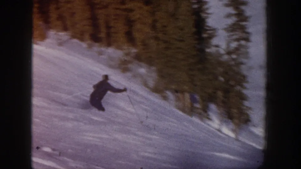 A lone skier continues downhill near evergreen woods in soft, grainy 8mm footage from Aspen.