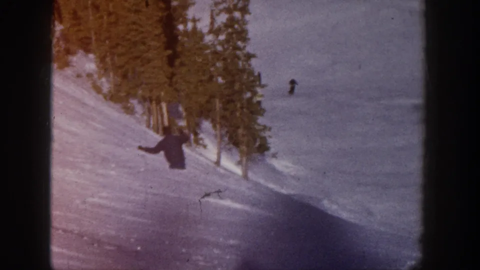 A distant skier crosses a wide snowy Aspen slope, framed by tall evergreens and dark 8mm gate borders.