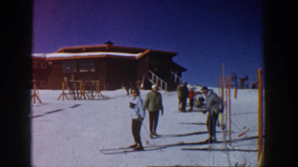 1961 Aspen resort scene with skiers gathered by racks outside a lodge, including one person adjusting gear in the foreground.