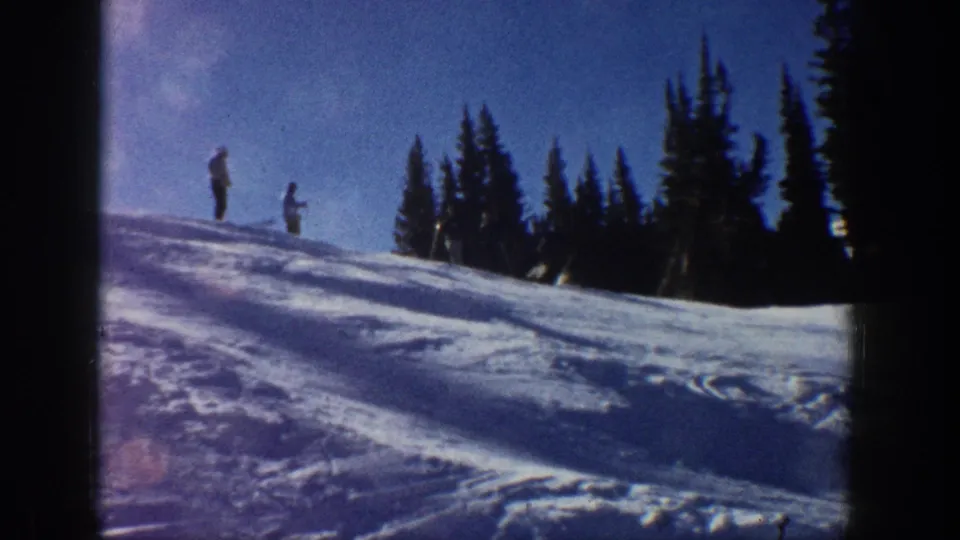 Two distant people stand on an Aspen snowy hillside in 1961 home-movie footage, with evergreen trees and blue sky behind them.