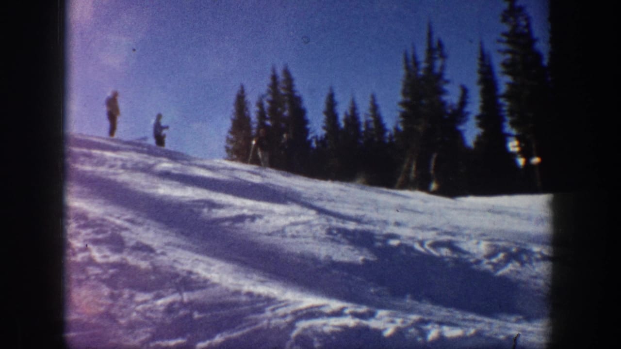 Aspen, Colorado home-movie frame from 1961 showing two skiers on a snowy hillside with evergreens and blue sky.