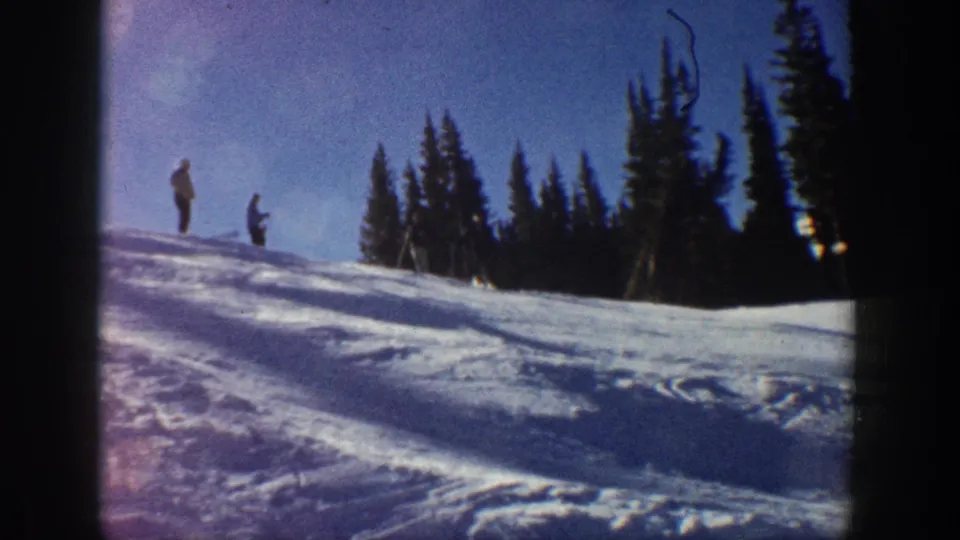 Aspen, Colorado 1961 home-movie still with two skiers waiting on a small snowy ridge near tall evergreens.