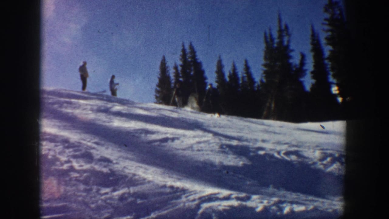 1961 Aspen home-movie frame showing two distant figures on a sunlit snowy slope near dark evergreen trees.