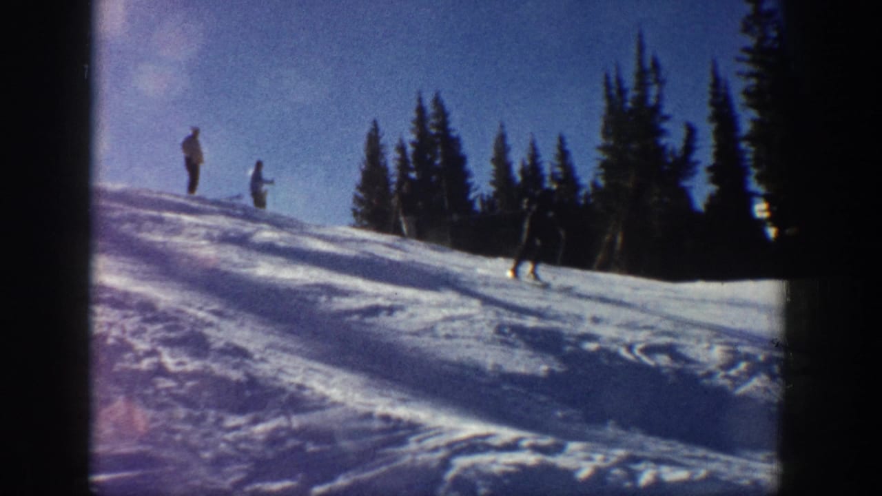 Distant skiers and standing figures appear on a snowy Aspen slope bordered by evergreen trees in 1961 footage.