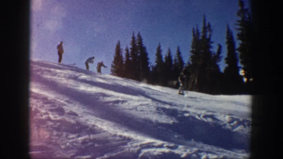 1961 Aspen footage frame with several distant figures on a snowy ski slope against a dense evergreen tree line.