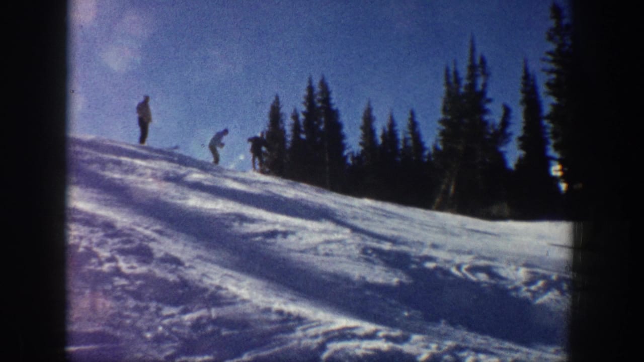 Aspen, Colorado 1961 home-movie frame showing distant skiers and standing figures on a bright snowy slope beneath a blue sky.