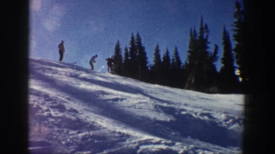 Aspen, Colorado 1961 home-movie frame showing distant skiers and standing figures on a bright snowy slope beneath a blue sky.