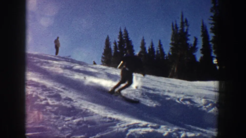 Aspen, Colorado 1961 home-movie frame showing a skier leaning into a turn on a sunlit snow slope with dark evergreens beyond.