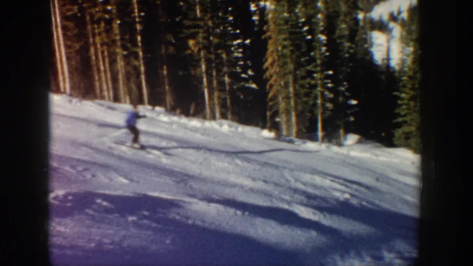 A lone skier crosses the lower-right frame on a snowy Aspen slope, with evergreen trees and visible 8mm film texture.